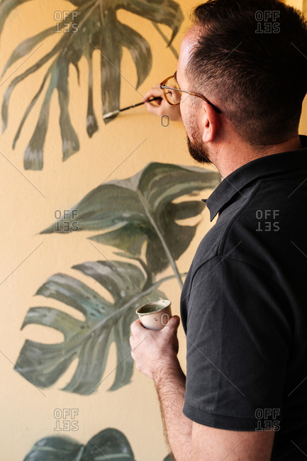 Side view of talented male artist painting green monstera leaves on wall in apartment