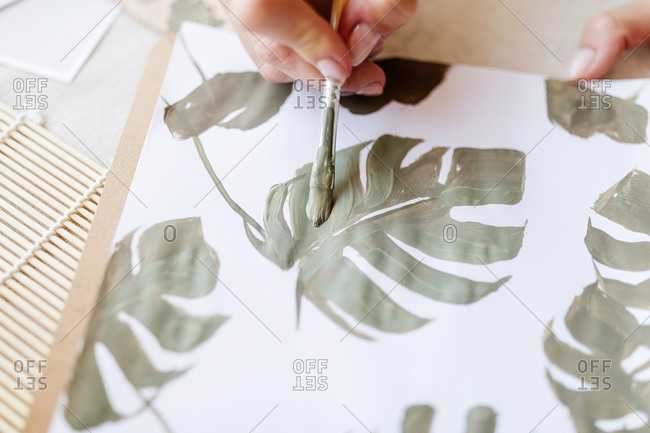 Unrecognizable crop female artist standing at table and painting green leaves of monstera plant on sheet of paper in art workshop