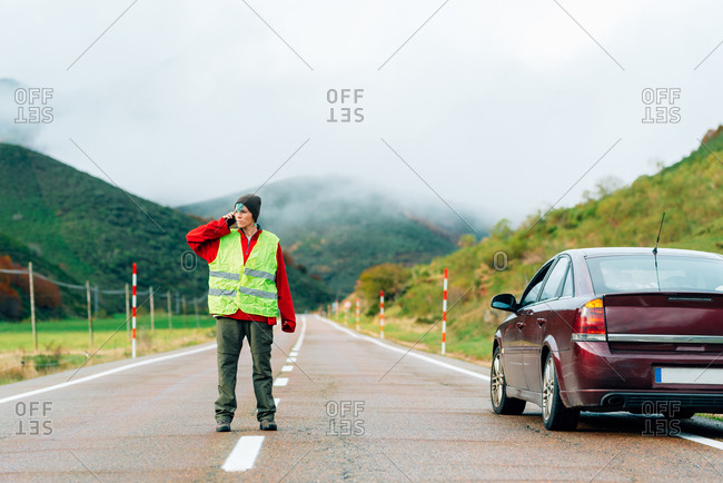 Female driver in reflective vest standing on road near broken car in mountainous area and talking on smartphone while calling for help