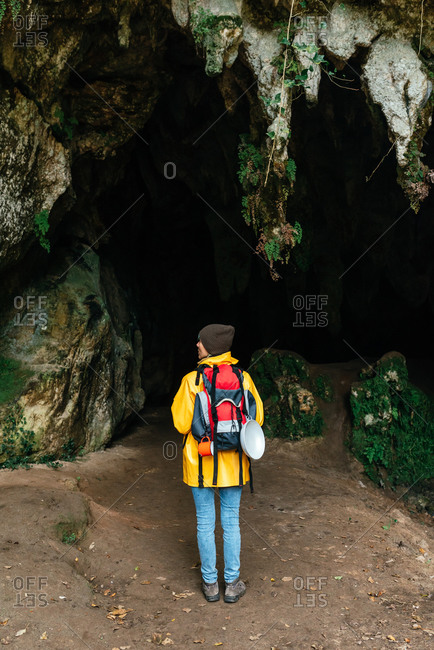 back view of female traveler with backpack and in outerwear standing in rocky arched passage near Cobijero Beach and looking at camera while enjoying autumn vacation in Asturias