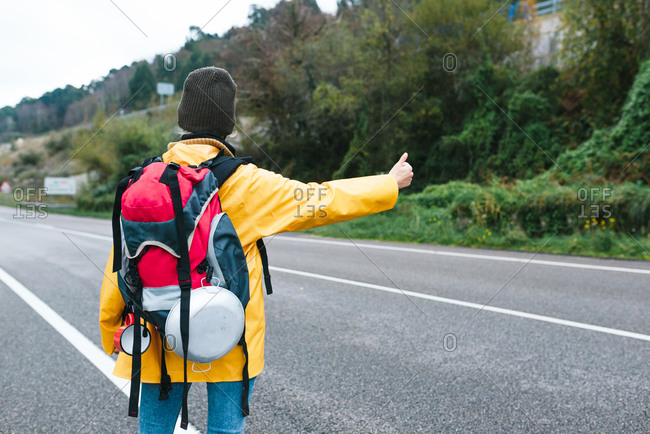 Back view of female backpacker in yellow raincoat standing with thumb up on roadside while hitchhiking in Asturias in autumn