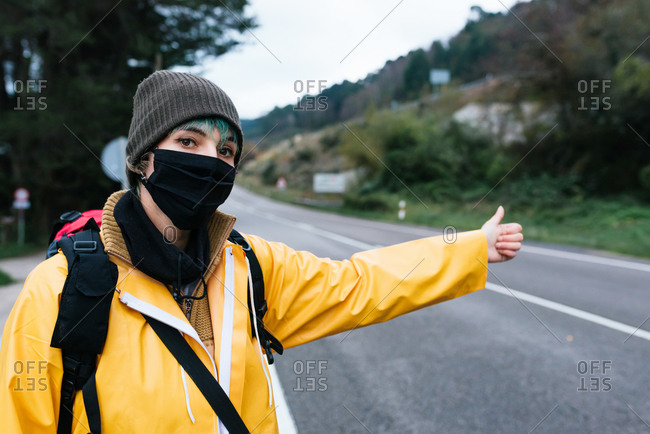 Female backpacker in yellow raincoat and protective mask standing with thumb up on roadside while hitchhiking in Asturias in autumn