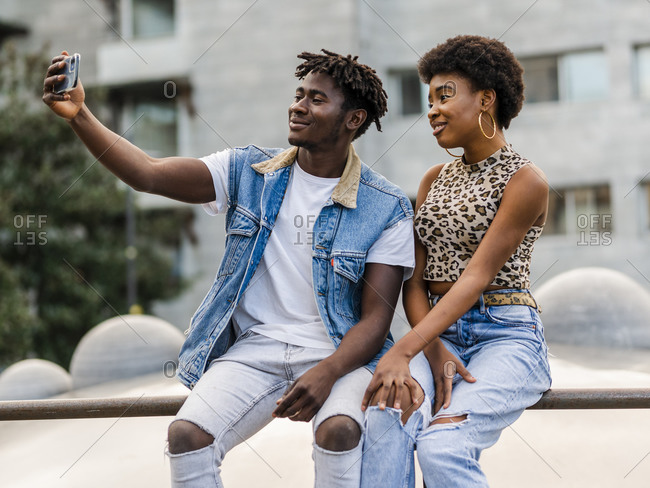 Cheerful young African American man and woman with curly hair dressed in trendy ripped jeans sitting on fence and taking selfie on smartphone while spending time together in city