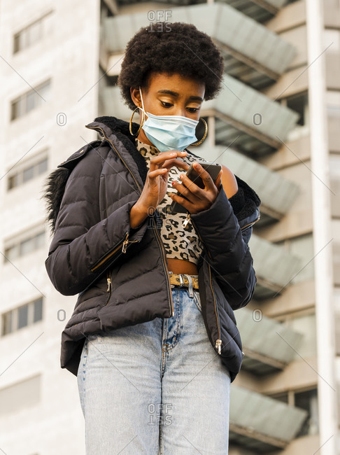 From below of modern young black female with Afro hairstyle wearing warm jacket and protective mask browsing mobile phone while standing near city building