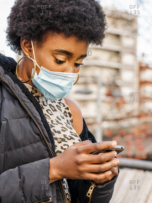 Modern young black female with Afro hairstyle wearing warm jacket and protective mask browsing mobile phone while standing near city building