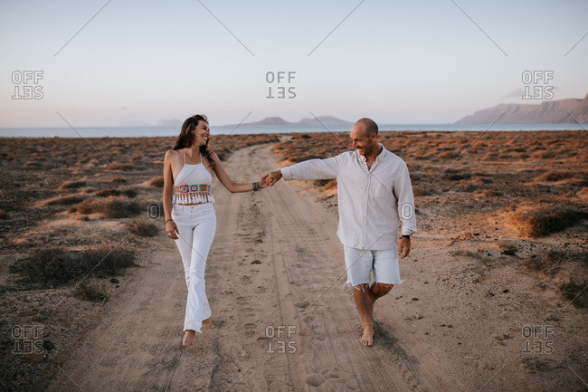 Delighted couple in white outfit holding hands and walking barefoot along sandy road in savanna at sunset