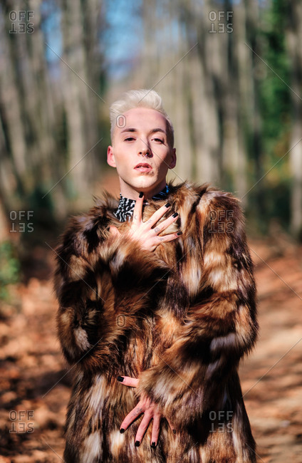 Confident young blond haired transgender male with makeup wearing stylish fur coat looking at camera while standing in sunny autumn forest