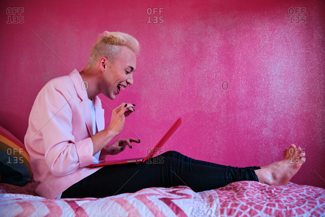 Side view of cheerful stylish focused young blond haired transgender male using red laptop while sitting in room with feminine pink colored interior at home
