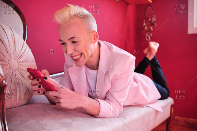 Happy stylish young blond haired transgender male with makeup browsing mobile phone while chilling on sofa in room with pink interior