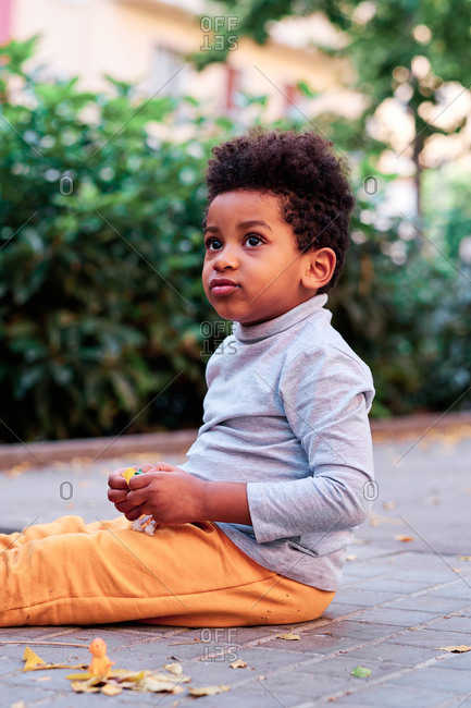 Side view of adorable black kid with Afro hairstyle sitting on pavement with toy and looking away