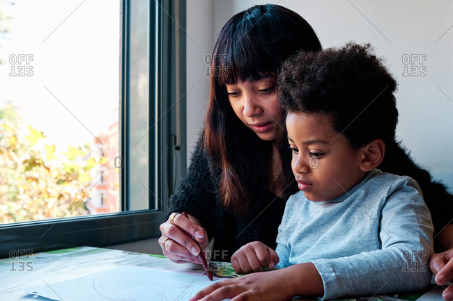 Caring ethnic mother sitting with curious child and drawing on paper together while enjoying weekend at home