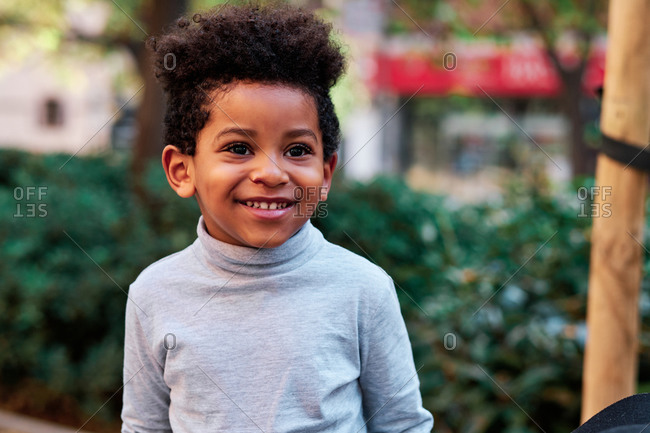 Cheerful cute African American child with curly hair standing on street in summer and looking away