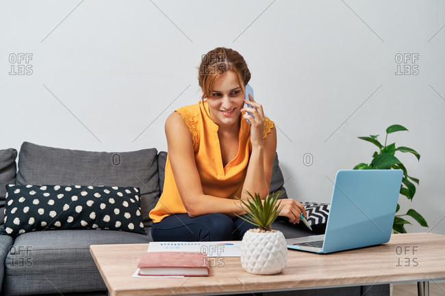 Content female entrepreneur sitting at table in living room and speaking on smartphone while discussing business ideas and using laptop