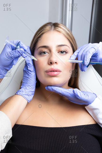 Graceful young female client with long blond hair sitting on medical chair while cut unrecognizable cosmetologists in latex gloves making rejuvenating facial injections procedure in beauty center