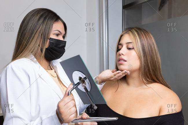 Unrecognizable female cosmetologist in medical mask and robe holding mirror and showing result of procedure to happy patient with long hair