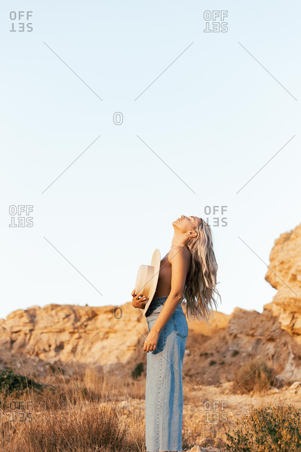 Side view of tranquil topless female in jeans standing in nature and covering breast with hat while enjoying sunset in summer