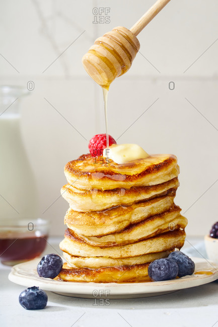 Pouring honey on a stack of fluffy breakfast pancakes with berries and butter