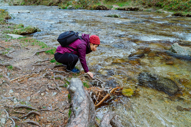 Side view of unrecognizable female adventurer in warm outfit resting on shore of narrow creek flowing between pine trees in autumn woodland in Sierra de Madrid in Spain
