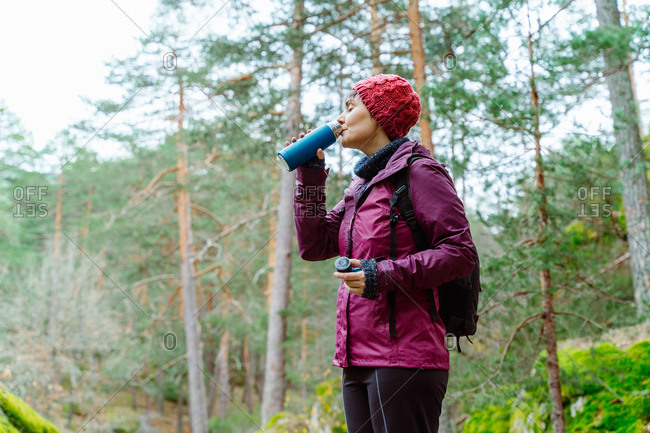 Side view of female adventurer in outerwear and with thermos standing near river with fast flow and enjoying nature during vacation in Sierra de Madrid