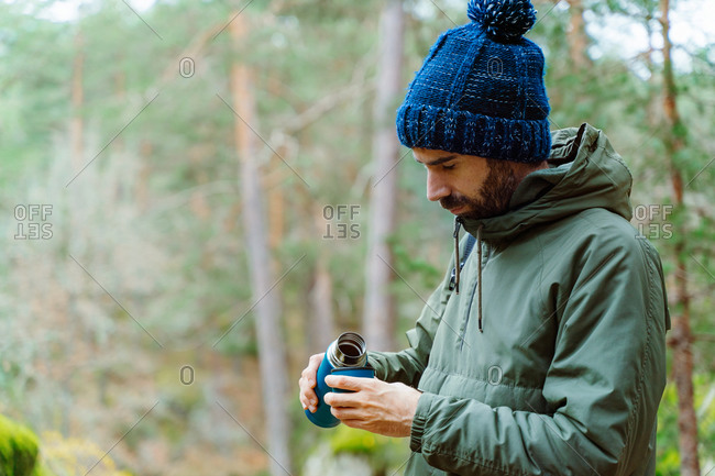 Side view of tranquil male traveler in warm clothes standing in woods and pouring aromatic hot tea in thermos mug during autumn vacation in Sierra de Madrid