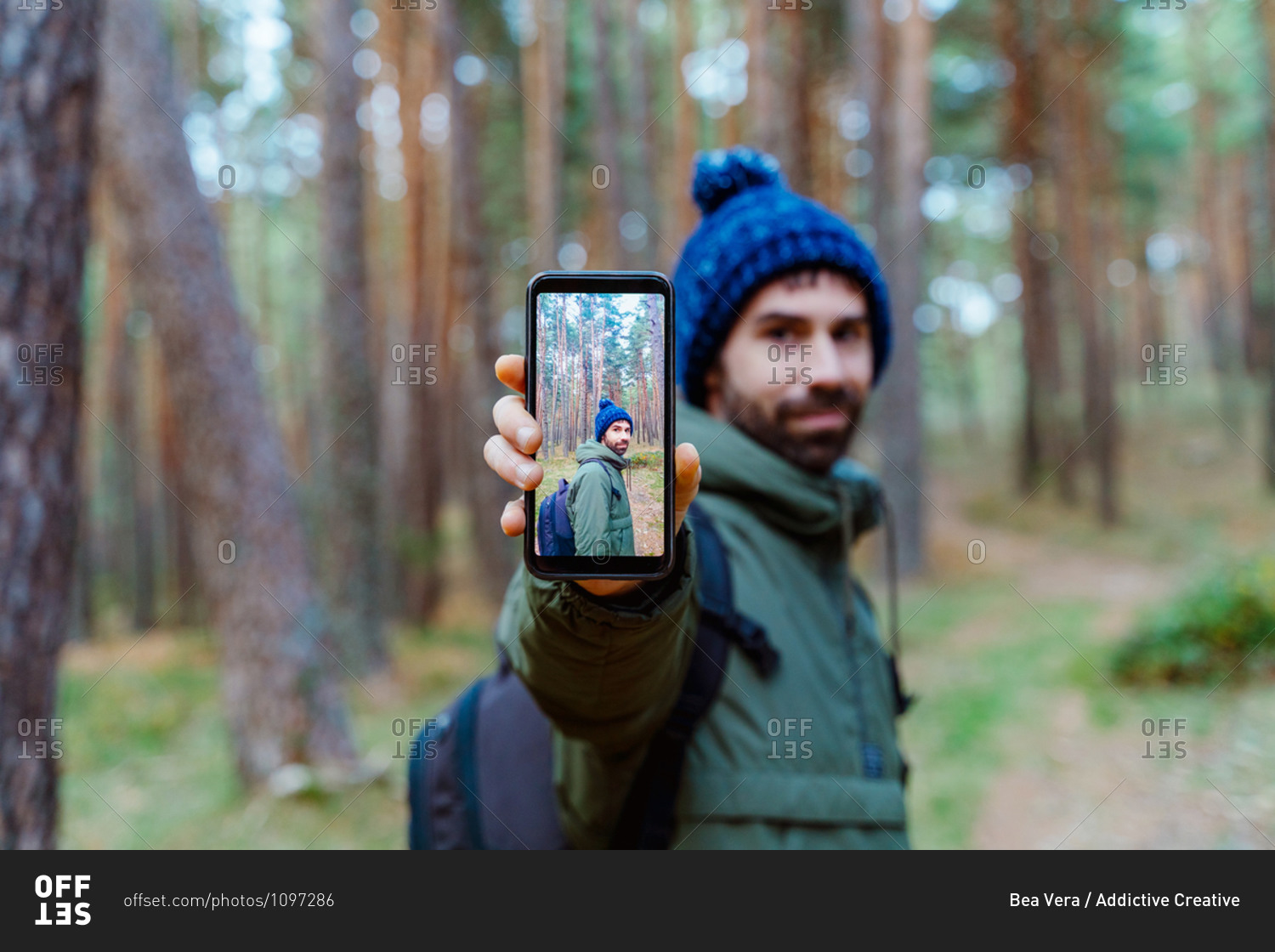 Side view of traveling man in warm outerwear standing in woods and showing smartphone with photo during autumn adventure in Sierra de Madrid