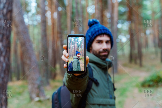 Side view of traveling man in warm outerwear standing in woods and showing smartphone with photo during autumn adventure in Sierra de Madrid