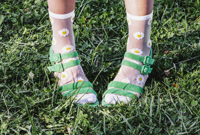 Legs of crop anonymous female wearing green sandals and socks with chamomile flowers print while standing on lawn in summer on sunny day