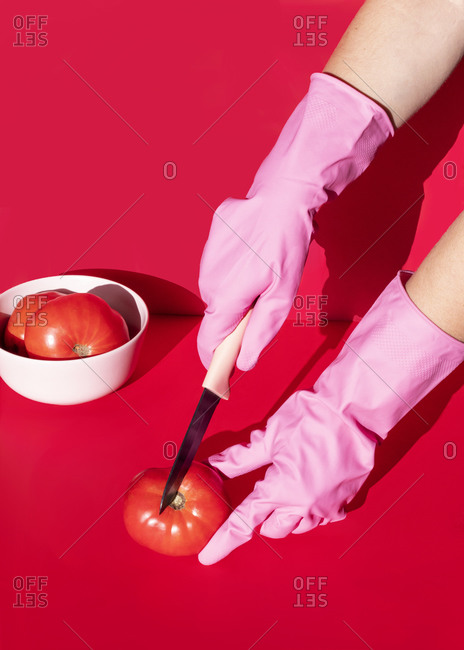 High angle of crop unrecognizable person in rubber gloves cutting ripe tomato on red background in studio