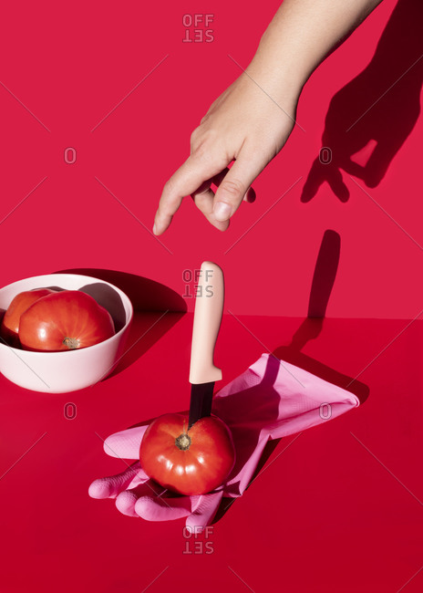 Cropped unrecognizable person hand near fresh tomato with sharp knife placed on rubber glove on red backwound in studio