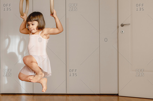 Adorable little girl in dress handing above floor on wooden gymnastic rings in bright room and looking away