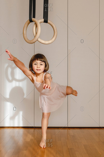 Adorable little girl in dress handing above floor on wooden gymnastic rings in bright room and looking away