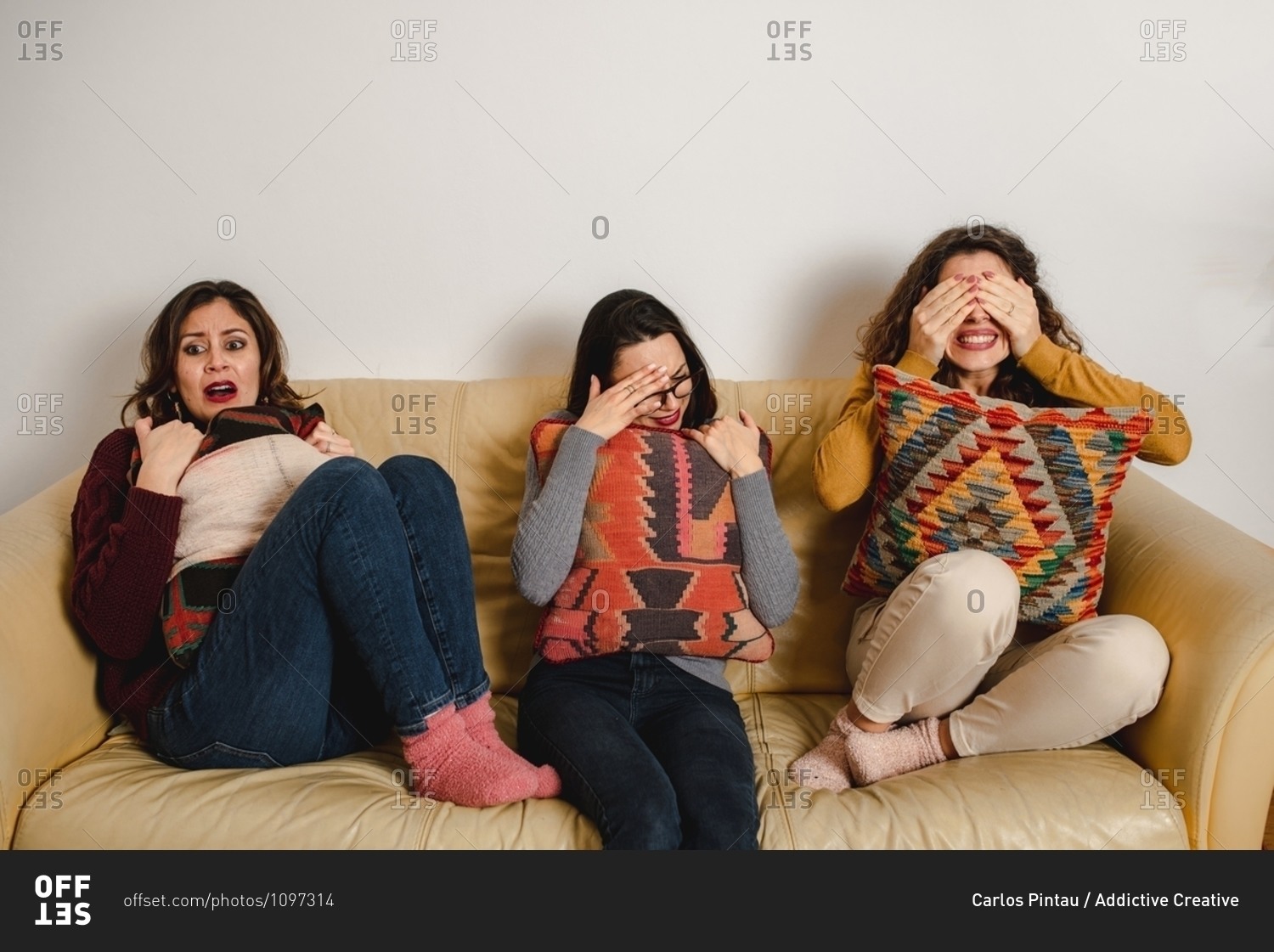 Group of frightened female friends sitting on sofa at home and watching horror movie together while covering eyes and feeling fear