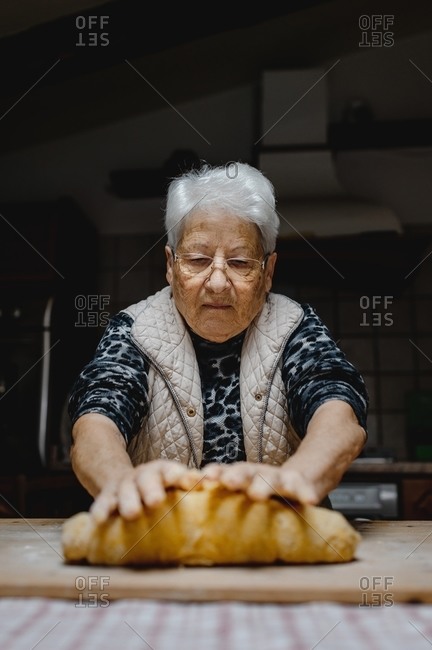 Senior female standing at table in cozy kitchen and kneading raw dough while preparing pastry for cooking domestic Italian tortellini