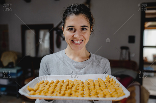 Delighted housewife standing in kitchen with baking pan full of homemade traditional tortellini and looking at camera