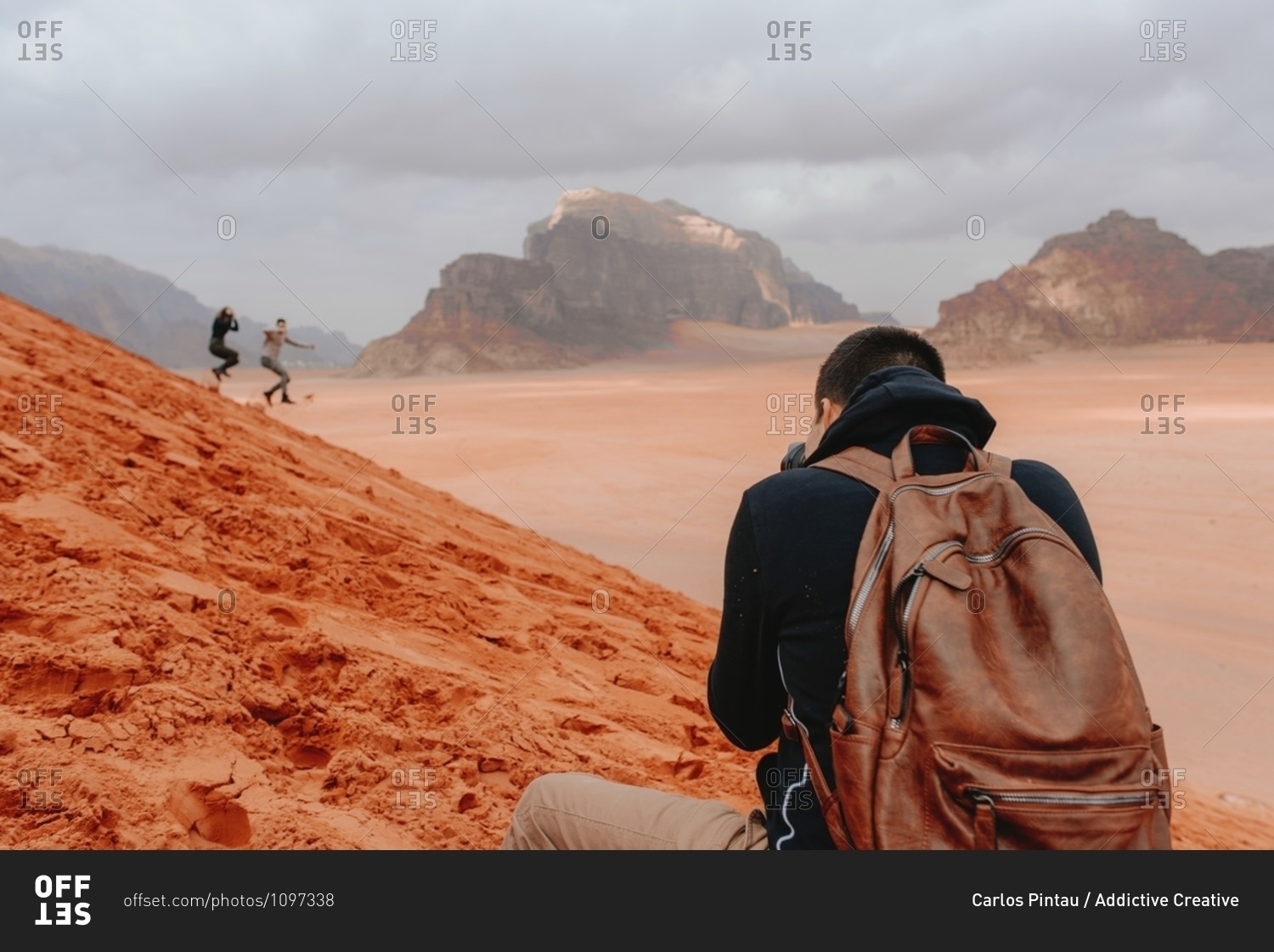 Back view of male traveling photographer taking pictures of people running down sandy hill in Wadi Rum sandstone valley during vacation in Jordan