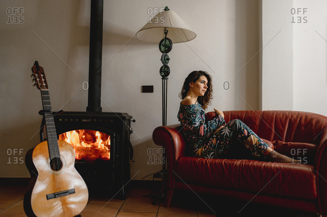 Attractive caucasian woman portrait sitting on the sofa. Girl wearing sleepwear next to the fireplace in the living room with a guitar. Cozy, comfortable, winter, domestic life concept.