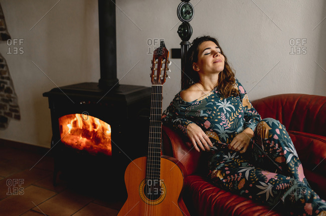 Close-up portrait of sensual attractive caucasian woman sitting on the sofa. Girl wearing sleepwear next to the fireplace with guitar in the living room. Cozy, winter, life at home concept.