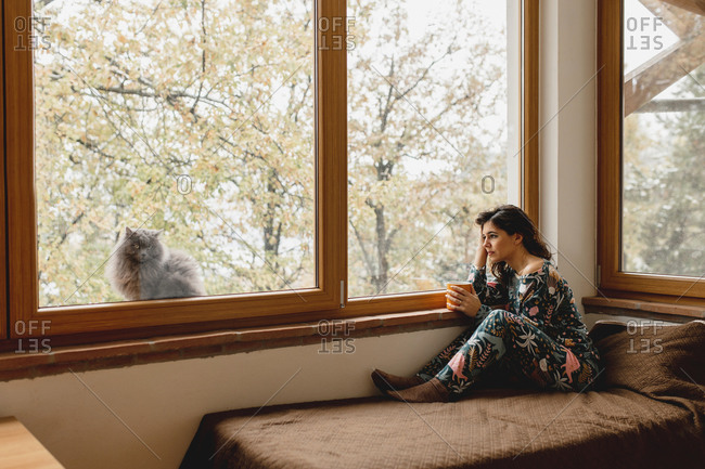 Sleepy, thoughtful woman sitting on the bed holding a cup of hot beverage. Cat looking at the camera through the glass window. Domestic life, relax, holiday concept.