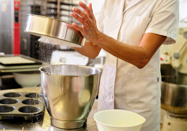 Cropped unrecognizable female chef in white uniform and hat sifting flour into metal bowl while preparing dough for pastry in bakery kitchen