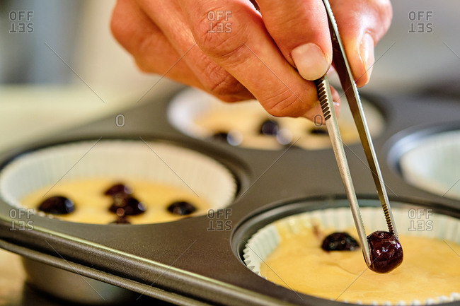 Closeup of crop anonymous chef with kitchen tongs putting berries into dough in muffing form while preparing pastry in kitchen