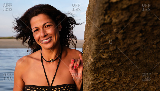Cheerful female in bikini standing near rock on Fuseta Beach and looking at camera while enjoying summer vacation at seaside in Algarve