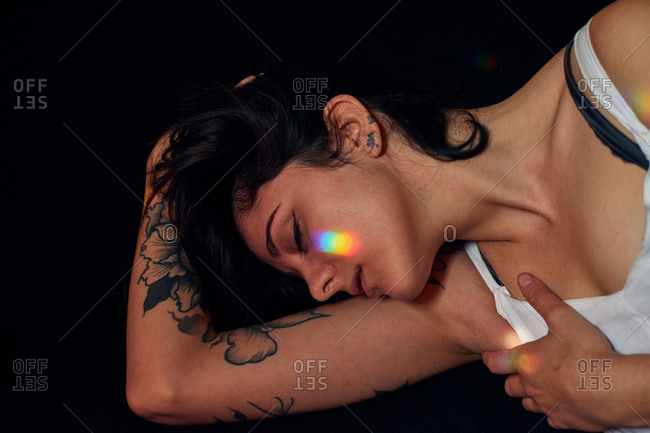 Serene female with tattoos lying on floor with closed eyes and  flares on faces on black background in studio
