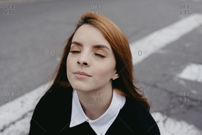 Young redhead female with wind in hair standing in city with eyes closed