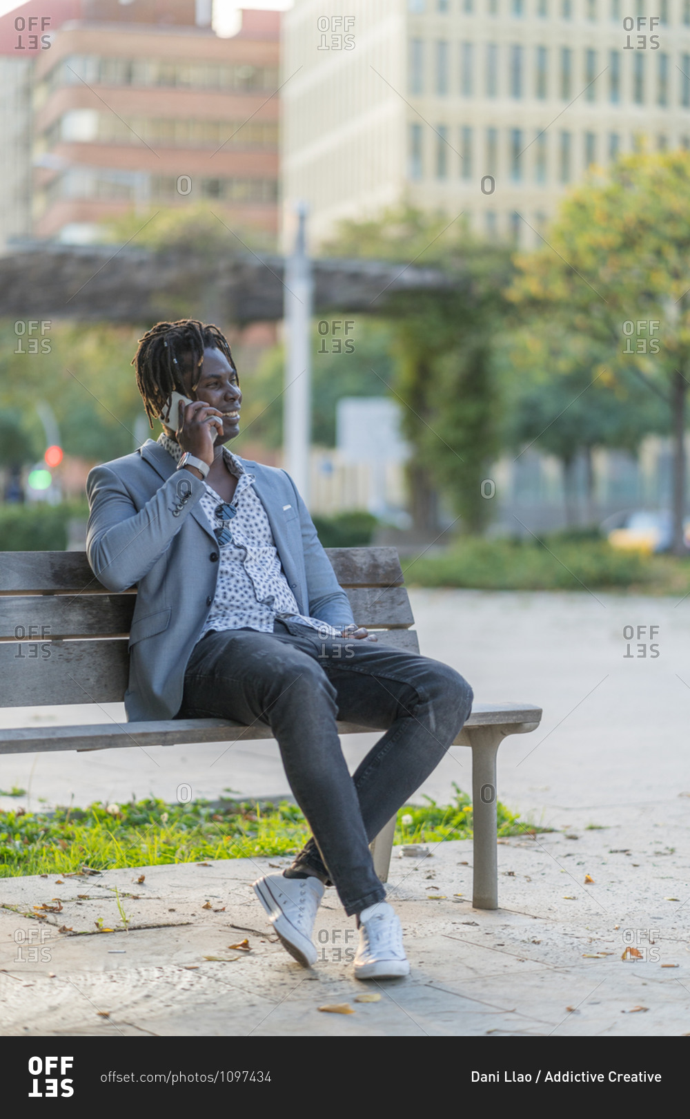 Cheerful African American male sitting on bench in city and talking smartphone while communicating on social media and relaxing at weekend