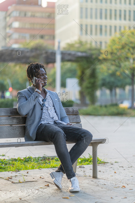 Cheerful African American male sitting on bench in city and talking smartphone while communicating on social media and relaxing at weekend