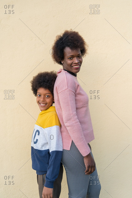 Side view of tender African American woman and boy standing back to back looking at camera on street in city against beige wall