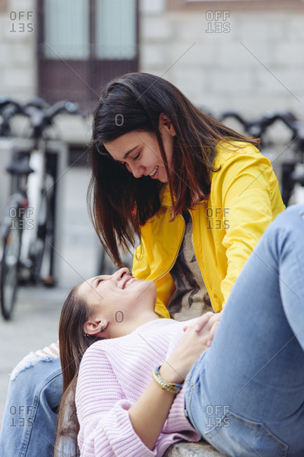 Gentle couple of lesbian females resting on stone bench on street while smiling and looking at each other
