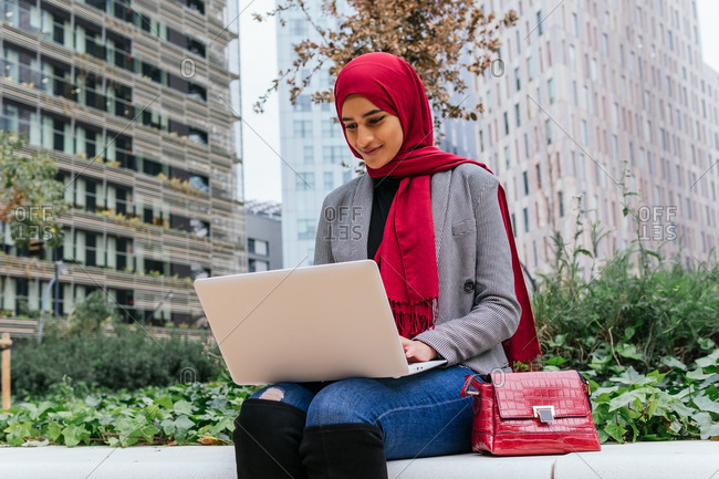 Busy Arab female entrepreneur in traditional headscarf sitting on street with laptop and working on new business project