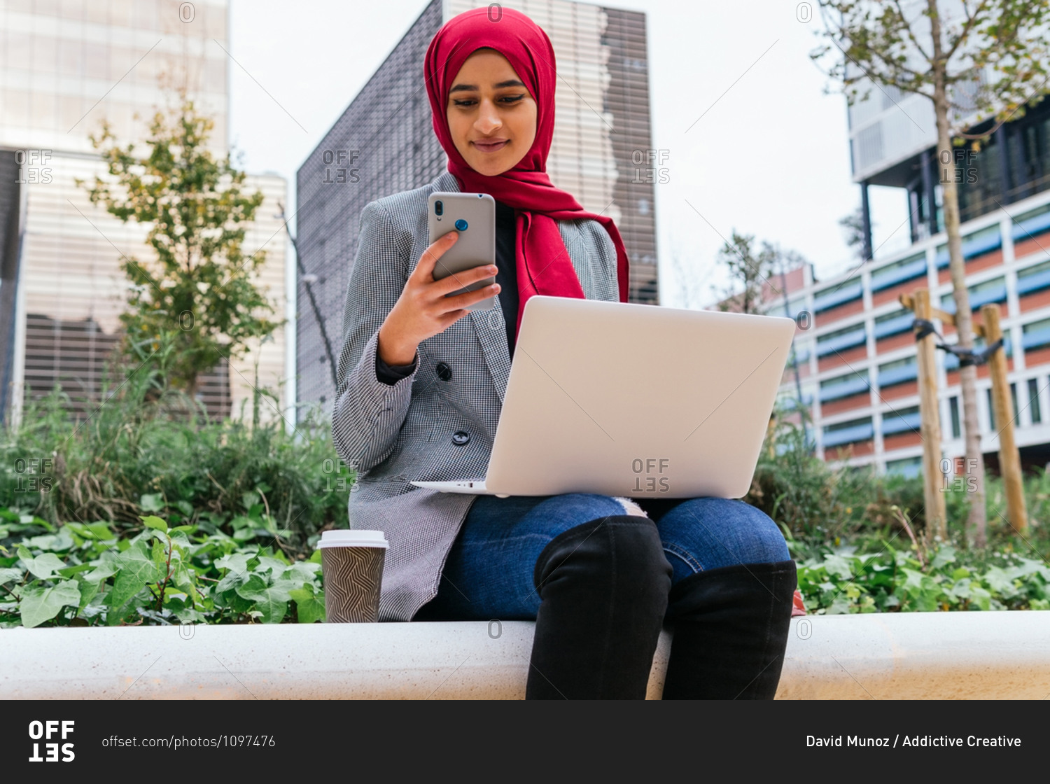 Cheerful Arab female freelancer in red hijab sitting with laptop on bench in city and browsing smartphone while working on startup project remotely