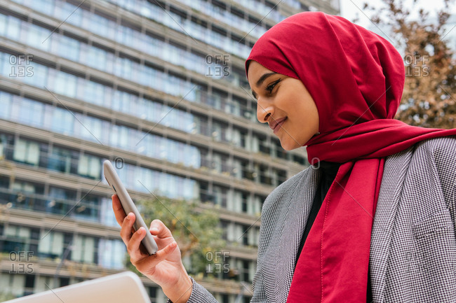 Cheerful Arab female freelancer in red hijab sitting with laptop on bench in city and browsing smartphone while working on startup project remotely